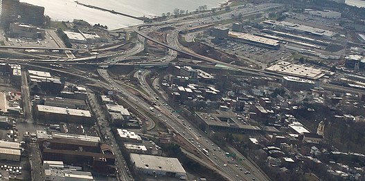 A series of highway ramps with multiple cars on them. A body of water is next to them, and they are surrounded by buildings.