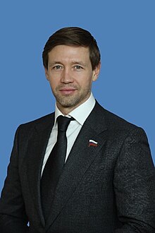 Colour photograph headshot of Artem Gennadyevich Sheikin wearing a black suit and tie, and a Russian flag pin badge, age 42