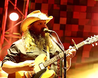 A man with long hair and a beard, wearing a cowboy hat, singing into a microphone and playing a guitar