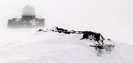 A NORAD Distant Early Warning Line (DEW) station in western Greenland is visible in the distance beyond the snow-drifted equipment pallets in the foreground of this photograph. The DEW Line was designed to track inbound ballistic missiles.