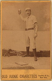 Бейсболист изображен стоящим в своей бейсбольной форме и снаряжении, используемом для кетчера.