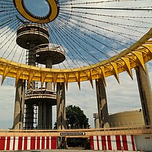 The interior of the Tent of Tomorrow, seen in 2017. At ground level is a red-and-white-striped wall topped by a yellow balcony. There is a passageway through the wall in the middle of the image. Above the wall, a set of concrete columns holds up two yellow girders. Wires extend from the girders to an elliptical ring in the middle. Three concrete observation towers are in the background.