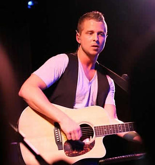 A photo of musician Ryan Tedder, wearing a black vest, performing with an acoustic guitar