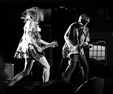 Une femme et un homme jouant de la guitare en performance.  La femme de gauche est vêtue d'une robe courte et l'homme de droite est en jean et chemise.