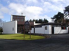 Modernistische Kirche mit weißem Putz mit großen modernen Buntglasfenstern und einem Holzkreuz, das aus dem Dach des zweistöckigen Turmelements des Gebäudes ragt