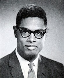 A dark haired man, wearing glasses and a suit and tie, looks into the camera