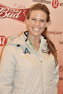 Photo of the torso and head of a female with a long brown ponytail who is smiling broadly. She is wearing a white nylon tracksuit with a green A on the right chest area. Behind her is an advertising background for Budweiser.