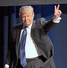 Donald Trump, dressed in a black suit with white shirt, blue tie, and black pants. He is facing toward the viewer, putting his left hand up in a "peace" symbol with his index and middle fingers raised. This is just after he spoke at the Conservative Political Action Conference in February 2011.