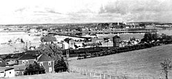 1930s bird's eye view of Bathurst, likely taken from College Sacre-Coeur. Looking east at railway in foreground, harbour and city in background.