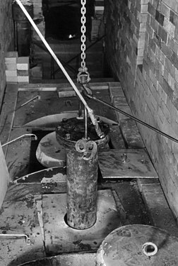 鐵鏈吊著一根柱形容器，容器一部分已沒入下方的設備中。