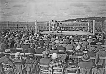 Photographie en noir et blanc de spectateurs assis autour d'un ring de boxe sur lequel deux boxeurs combattent.