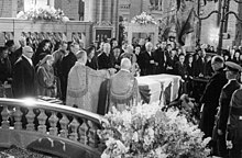Photographie en noir et blanc montrant deux prélats et une foule d'homme et de femmes en tenues de deuil entourant un cercueil dans une égalise.