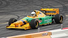 Foto de um monoposto de Fórmula 1 amarelo e verde, vista de três quartos, em uma pista de corrida