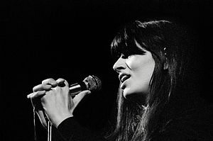 Fotografia em preto e branco de uma mulher com longos cabelos escuros segurando um microfone e cantando