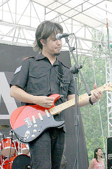 An Indonesian man with long hair, playing a red guitar. His face is partially obscured by a microphone.