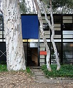 Aperçu de la façade d’une maison contemporaine. Cadres géométriques sombres. Panneaux colorés et vitrages. Au premier plans, des troncs d’arbres.