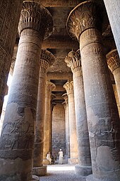 Habitación grande y en penumbra llena de columnas altas y gruesas.  Los capiteles de las columnas tienen forma de flores estilizadas.