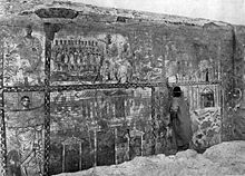 Photo en noir et blanc. Un homme se tient devant un mur recouvert de symboles.