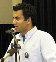 A man giving a speech. He wears a white blouse with a dark label pin. In front of him, there are two microphones.