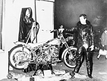 Director Kenneth Anger on set with a motorcycle and a skeleton dressed as the Grim Reaper Kenneth Anger (Scorpio Rising).jpg