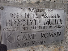 Ciemna metalowa tablica zapieczętowana w skale z komentarzem: „19 listopada 1911 – Montaż kładki Hyppolyte Müller – Société des alpinistes Dauphinois – Rzymski obóz 40 minut drogi”