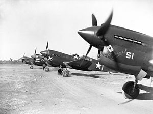 Quatre avions de chasse à moteur à piston Curtiss P-40 monoplace en livrée sombre s'alignent sur une piste d'atterrissage en terre battue