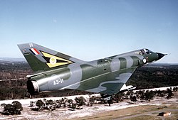 A close up view of a flying jet. The aircraft is painted in a military camouflage pattern, and a tiger's head symbol is painted on the tail fin. The back of the pilot can be seen in the cockpit, along with trees spread out below.