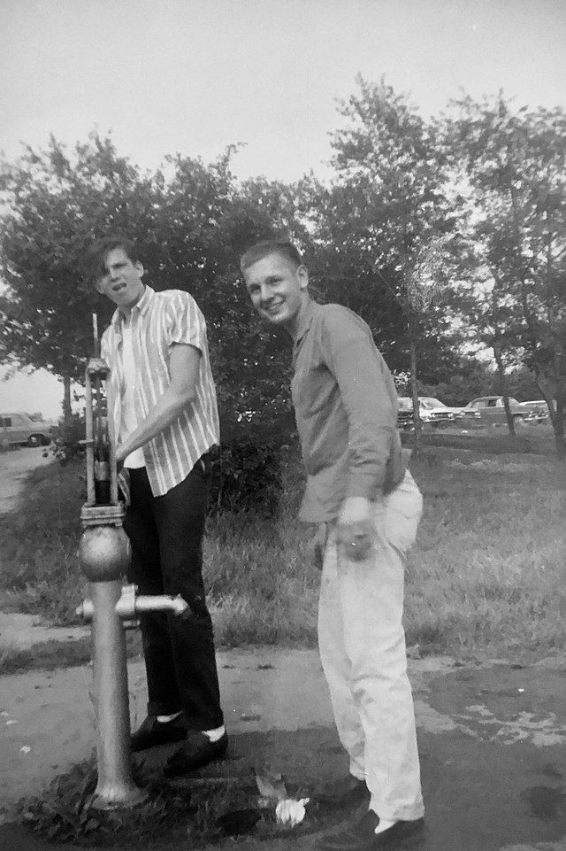 Black-and-white photo of two young men standing by a simple hand-operated water pump, with trees in the background
