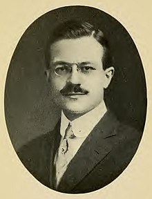 Une photo de portrait d'un homme blanc avec des lunettes pince-nez et une moustache, portant un manteau et une cravate, faisant face et regardant la caméra.