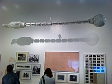 maquette d'une long vaisseau spatial tubulaire suspendu dans une salle d'exposition au dessus de photos de tournage.