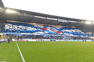 Toute la hauteur d'une tribune rectiligne est couverte par un écusson circulaire, à l'effigie du RC Strasbourg, porté au-dessus d'eux par des supporters. Autour, des cartons colorés tenus par le public dessinent des lignes convergentes.