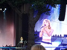 Taylor Swift standing near a fake tree, with a screen showing her face in the background