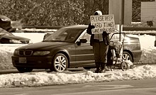 A woman holding a sign by a road, writing "Please help hard time, unemployed" in Massachusetts Unemployed.jpg