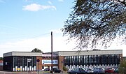 A building approximately 3 stories high. For the most part, the building is made up of horizontal columns of an opaque white panel up to around head height, with 3 windows arranged vertically above this. The columns are approximately 0.5m (1.5ft) wide and run from just below the roof to just above ground level and are separated by vertical wooden beams. Above the columns of windows a corrugated white border runs along the top of the building.