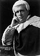 A photographic portrait of a man, showing his head and shoulders; he is learning on one hand and has his elbow propped up on a desk. He is wearing an English Judge's wig and robes.