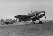 A black-and-white photograph of a twin-engine fighter aircraft standing on a grass field, shown in profile, viewed from the right. The aircraft is appears grey, with a teethed mouth painted on the nose. The twin rudder at the rear bears a black swastika. Further decorations include lines, black and white crosses on the body and on bottom of the wing; a number M8 is visible. The landing gear is extracted and the propellers are turning.