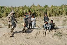 Photo en couleurs d'un soldat en uniforme de combat tenant un fusil à la main marchant devant un groupe d'enfants sur un terrain de sable avec une fôrêt en arrière-plan