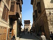 Several people walk down a small empty lane overshadowed on both sides by three-story buildings with shrouded balconies and windows of Islamic style