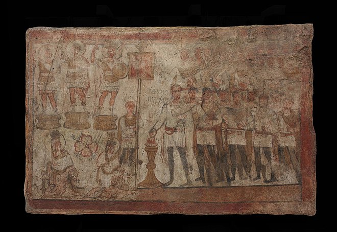 Julius Terentius Performing a Sacrifice. Commissioned by the Roman tribune at Dura-Europos, Julius Terentius (depicted and identified here by Latin inscription), this painting mixes Greek, Roman, and Palmyrene elements, perhaps as a gesture of diplomacy. Performing an official sacrifice in front of a military standard, Terentius stands with his men. Although in Roman military attire, these soldiers are Palmyrenes; one of them (Themes, son of Mokimos) is identified in Greek as a priest. The sacrifice, a burning of incense, was an act of worship common in both Near Eastern and Roman sanctuaries. Here it takes place in the presence of divinities, all nimbate. The Tychai of Dura and Palmyra are shown in Hellenistic style incorporating some Near Eastern features. Three male statues of deities enigmatically combine Roman cuirasses and Palmyrene attributes (such as the peaked helmet). Their lack of identifying labels creates ambiguity as to whether they represent Palmyrene gods or deified Roman emperors. This may have been done intentionally, to appeal to viewers of different backgrounds. Julius Terentius Performing a Sacrifice (Dura Europos).jpg