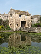 Un bâtiment en pierre de deux étages avec une arche véhiculaire au centre.  Il a un toit en pierre et les pignons à chaque extrémité ont des fleurons en boule de pierre.  Devant la guérite se trouve un pont de pierre traversant un large fossé sur lequel poussent des plantes aquatiques.