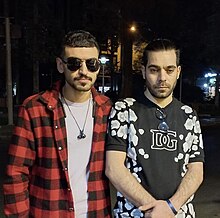 From left to right, Matin Mannani and Ali Tahoune standing side by side. Matin is wearing sunglasses and a necklace, over a white t-shirt with a plaid black and red button up over the top. Ali is wearing a black top with a floral print and DG in the middle. Ali has glasses hanging on the collar of his shirt.