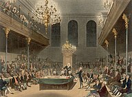 View of a large room, showing the Speaker of the House sitting at the end. Down each side of the room, MPs are sitting; one MP is standing on the right, giving a speech. Balconies are on either side, with spectators visible.