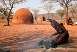 Himba woman prepares a fire. Himba huts in the background.