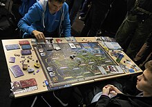 Fotografia colorida de duas pessoas sentadas em uma mesa jogando um jogo de tabuleiro que inclui cartas e dados.