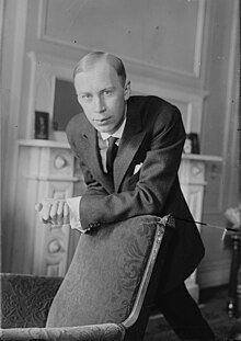 Black and white photo of Prokofiev standing besides a fireplace, his arm rested on the mantlepiece