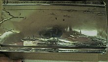 Tapa de plata de la caja-estuche de brezo conmemorativa de la participación en la batalla aeronaval de Punta Stilo.  En el borde superior los detalles históricos, en el borde inferior la descripción de los eventos a bordo.