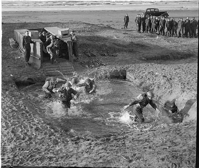 Commandos simulate an amphibious landing by disembarking from a dummy landing craft into a shallow pit filled with water.