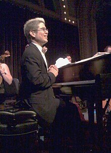 A smiling man with glasses sits at a piano on stage