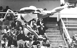 A man in an athletic uniform is diving forward, face-down over a high jump bar. His right leg is extended straight, parallel to the bar, while his left is bent at the knee and is just touching the bar as he leaps over it.