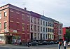 A row of four three-story flat-roofed brick buildings in various colors seen from across a corner. There are people standing on the other corners. The building nearest the camera, at the left, has a sign at street level saying 